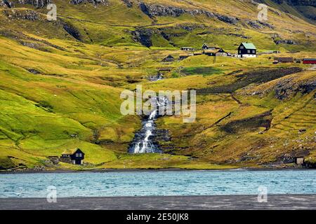 Kleiner Wasserfall und kleine Häuser im grünen Tal. Küste des Atlantischen Ozeans. Ebbe. Herbst im kleinen Dorf Saksun. Streymoy, Färöer. Stockfoto