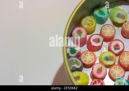 Bunte Lutscher in verschiedenen Geschmacksrichtungen in einer Blechdose auf einem weißen Tisch, Platz für Text. Süßigkeiten mit der Zeichnung der Früchte für die Kinder Stockfoto