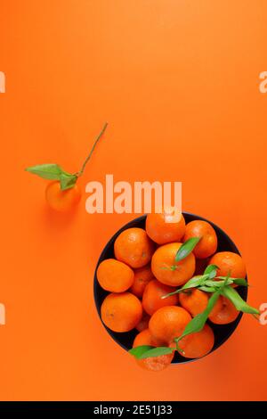 Mandarine mit Blättern und schwarzer Schale mit Mandarinen auf Orange Hintergrund Stockfoto