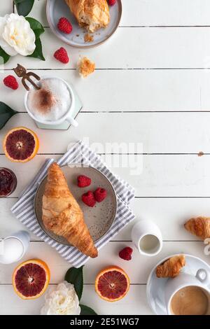 Flache Verlegung eines Frühstückstisches mit Croissants mit Marmelade, Kaffee, Himbeeren, Blutorange, gekochtem Ei und Blumen auf einem weißen Holztisch, vertikal Stockfoto