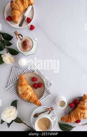 Flacher Frühstückstisch mit Croissants, Kaffee, Himbeeren, Blutorange, gekochtem Ei und Blumen auf einem weißen Holztisch, auf weißer Marmorbrandung Stockfoto