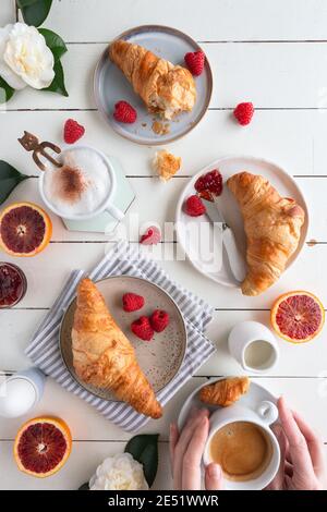 Die flache Lage des Frühstückstisches mit den Croissants mit der Marmelade, dem Kaffee, der von den weiblichen Händen gehalten wird, den Himbeeren, der Blutorange, dem gekochten Ei und den Blumen auf dem weißen Holz Stockfoto