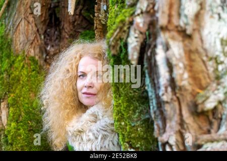 Eine rothaarige Frau in den Fünfzigern mit Pelzkragen blickt zwischen zwei moosbedeckten Baumstämmen, Kopierraum Stockfoto