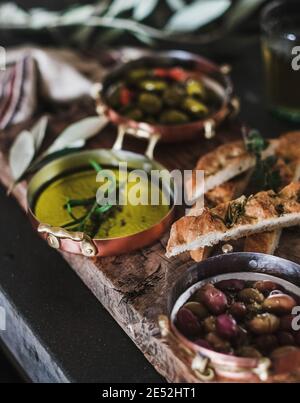 Eingelegte griechische Oliven, Olivenöl in Kupfergefäßen und Herbed Focaccia Scheiben auf rustikalem Holzbrett, selektiver Fokus, Nahaufnahme. Traditionelle mediterrane Meze Vorspeisen Platte Stockfoto