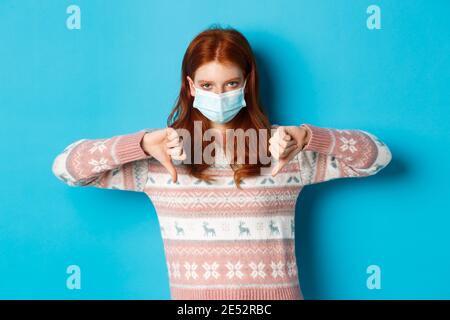 Winter, covid-19 und Pandemiekonzept. Aufgeregt und wütend Rotschopf Mädchen in Gesichtsmaske zeigt Missbilligung, Daumen nach unten in Abneigung, über blau stehen Stockfoto