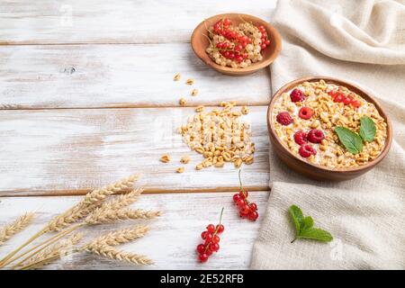 Weizenflocken Haferbrei mit Milch, Himbeere und Johannisbeere in Holzschale auf weißem Holzhintergrund und Leinentextilien. Seitenansicht, Kopierbereich. Stockfoto