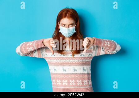 Winter, covid-19 und Pandemiekonzept. Moody Rotschopf Mädchen in Gesichtsmaske zeigt Daumen nach unten, missbilligen und nicht mögen, stehen vor blauem Hintergrund Stockfoto