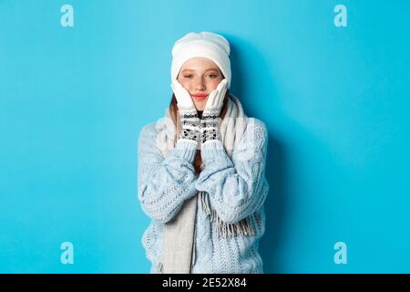 Winter- und Urlaubskonzept. Nettes Mädchen in weißen Beanie, Pullover und Handschuhe, quetschen Wangen und lächeln zufrieden, warm-up nach Kälte im Freien Stockfoto