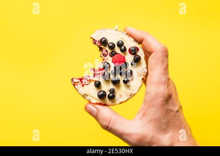 Hand halten knusprig aufgeblasen Reiskuchen mit frischen Früchten Blaubeeren Himbeeren vor dem gelben Hintergrund - gesund Bio-Vegetarier oder ve Stockfoto