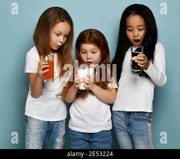 Drei überraschte Kinder Mädchen Freundinnen in weißen T-Shirts stehen und schauen auf Gläser Wasser, Milch und frischen Saft Stockfoto