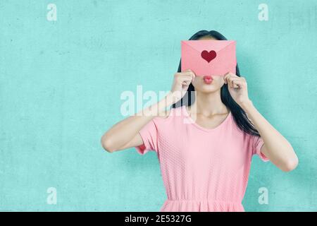 Asiatische Frau hält einen rosa Umschlag mit Herz mit einer blauen Wand Hintergrund. Valentinstag Stockfoto