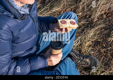 Ein Mann sitzt im Herbst auf dem Gras und isst ein Käse- und Wurstbrot und trinkt Kaffee. Ein schneller und leckerer Snack beim Spaziergang an der frischen Luft. Stockfoto