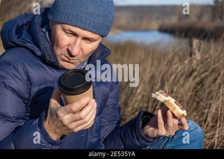 Im Herbst sitzt ein Mann auf dem Gras, isst ein Sandwich mit Käse und Wurst und schaut auf ein Glas Kaffee. Ein schneller und leckerer Snack beim Wandern. Stockfoto