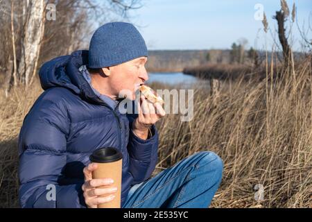 Ein Mann sitzt im Herbst auf dem Gras und isst ein Käse- und Wurstbrot und trinkt Kaffee. Ein schneller und leckerer Snack beim Spaziergang an der frischen Luft. Stockfoto