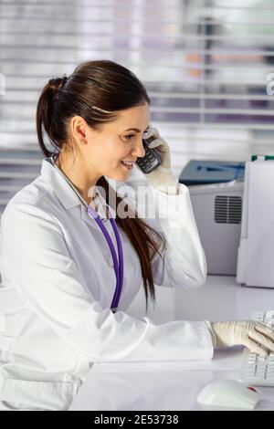 Frau Arzt, die Patienten am Telefon beraten, haben ein nettes Gespräch, sitzen am Schreibtisch am Arbeitsplatz, tragen weißen medizinischen Anzug und Blick auf Bildschirm von comput Stockfoto