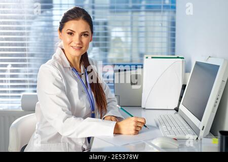 Doctor Frau sitzt am Schreibtisch Notizen machen, am Arbeitsplatz, in medizinischen Anzug. Professionelle Therapeut Blick auf Kamera und lächeln Stockfoto