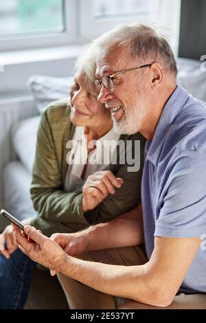 Ältere kaukasische Paar zu Hause mit Smartphone, sie sitzen zusammen, Ruhe, lächelnd Stockfoto