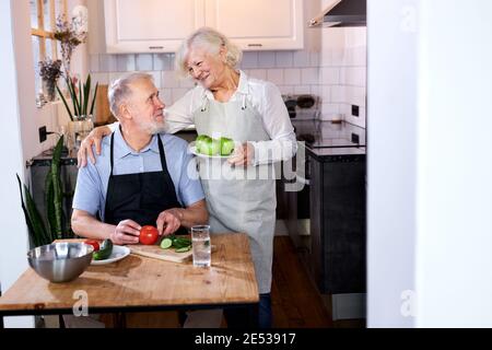 Ältere Paar in der Küche zu Hause, Senior Mann in Schürze sitzt schnitzen frisches Gemüse, grauhaarige Frau hält Äpfel in den Händen und spricht mit h Stockfoto