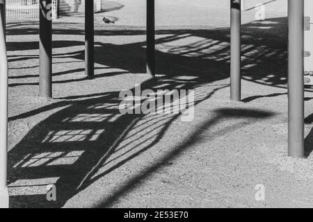 Schatten einer aufrechten Treppe und Geländer fallen zwischen die Metallstangen, die sie stützen, und werden auf das Gras eines Kinderspielplatzes geworfen. Stockfoto
