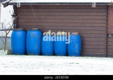 Fünf blaue Fässer in verschiedenen Größen stehen vor einem Holzhaus. Es ist Winter und es gibt Schnee auf der Wiese. Stockfoto