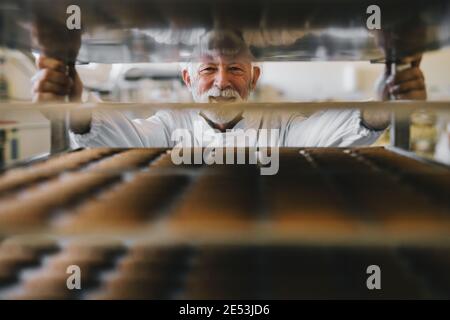 Bild von professionellen reifen männlichen Bäcker Mann in weiß Arbeit Uniform Blick auf die Kamera. Stehen vor den Regalen voll mit frisch gebackenen Koch Stockfoto