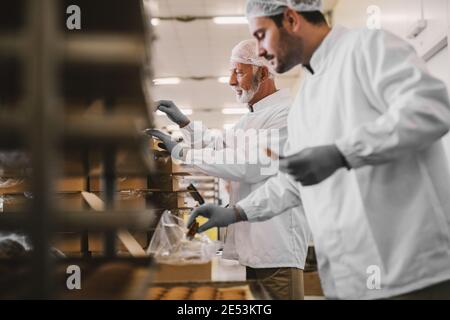 Bild von zwei männlichen Mitarbeitern in der Lebensmittelfabrik in steriler Kleidung, die frisch zubereitete Kekse verpackt haben. Im hellen Raum stehen. Stockfoto