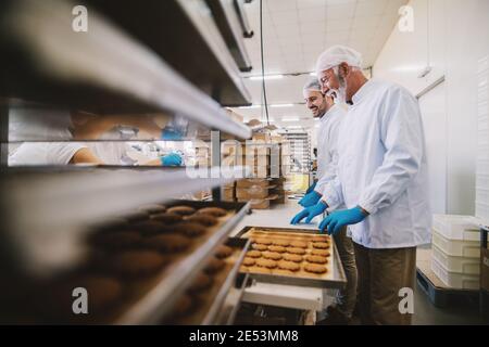 Bild von zwei männlichen Mitarbeitern in der Lebensmittelfabrik in steriler Kleidung, die frisch zubereitete Kekse verpackt haben. Im hellen Raum stehen. Stockfoto