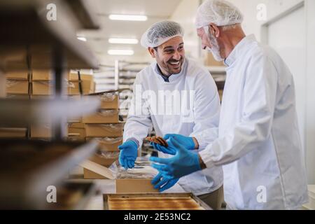 Bild von zwei männlichen Mitarbeitern in der Lebensmittelfabrik in steriler Kleidung, die frisch gebackene Kekse verpackt und Spaß hat. Stockfoto