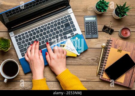 Mädchen hält Debitkarte, wählt Geschenke, macht Kauf, Laptop, Kaffeetasse, zwei Herzen, Tasche auf Holztisch Draufsicht Flat Lay Holiday Einkaufsliste Stockfoto