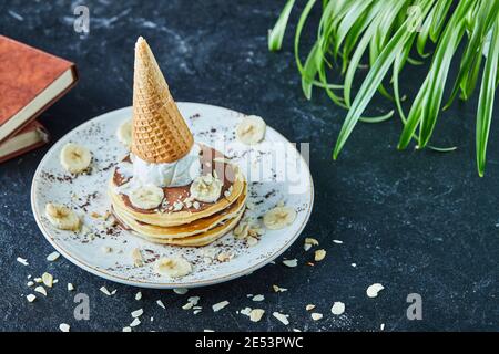 Pfannkuchen mit Eiskegel, Bananen, Kakaopulver und Bücher auf der weißen Platte auf dem dunklen Hintergrund Stockfoto