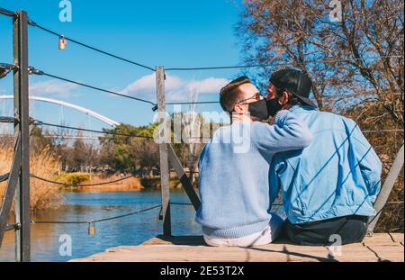 Junge Valentines Gay Paar Küssen Einander Tragen Schützende Gesicht Masken sitzen in hölzernen Bridg Stockfoto