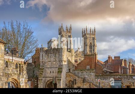 Die nach Westen gerichteten Türme des York Minster erheben sich über die Dächer einiger Häuser. Ein altes Stadttor ist im Vordergrund und ein wolkenvoller Himmel ist abo Stockfoto