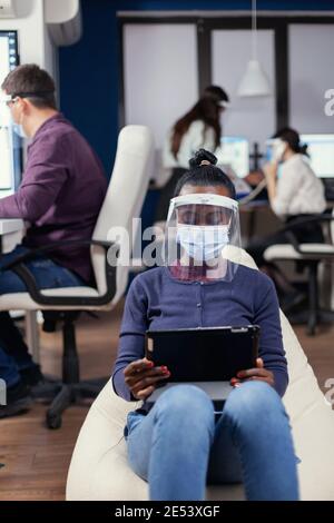 Afrikanischer Manager Notizen auf Tablet-pc trägt Gesichtsmaske gegen covid19 am Arbeitsplatz. Multiethnisches Geschäftsteam, das während der globalen Pandemie mit Coronavirus soziale Distanz respektiert. Stockfoto