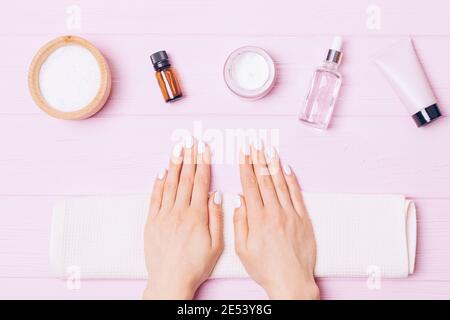 Weibliche Hände mit weißer Maniküre neben kosmetischen Produkten für Nagelpflege, Nagelhautöl, Creme und Lotion auf rosa Hintergrund, Draufsicht. Stockfoto