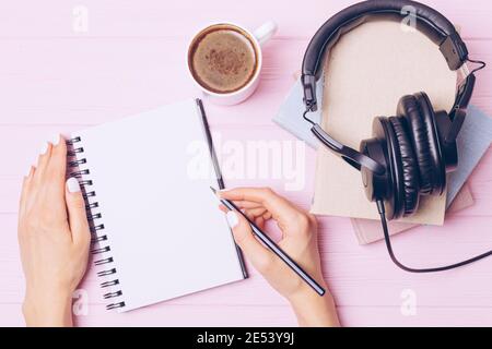 Weibliche Hände schreiben in leerem Notizbuch mit Bleistift neben Büchern, Kopfhörern und einer Tasse Kaffee, Draufsicht. Stockfoto