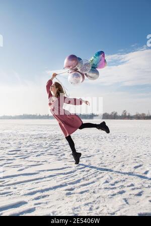 Foto in voller Größe von zarten anmutigen Teenager-Mädchen hält viele bunte Ballons in ihren Händen. Stehen im Schnee in einem rosa Kleid, ein sonniger Tag. Moment Stockfoto