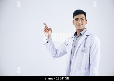Lächelnd positiver junger indischer Arzt im Labcoat, der weg zeigt, isoliert auf Weiß Stockfoto