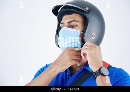 Junge seriöse indische Kurier in Schutzmaske Befestigung helment und Sich bereit machen, dem Kunden Fuß zu liefern Stockfoto