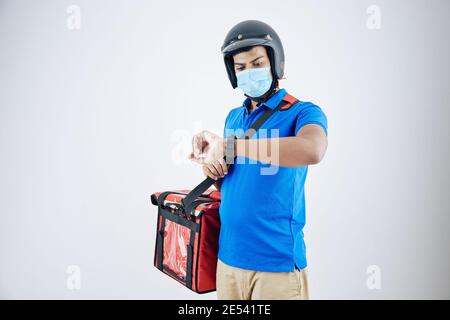 Junge ernsthafte Kurier in Helm und medizinische Maske trägt Kühler Tasche und Kontrollzeit auf Smartwatch Stockfoto