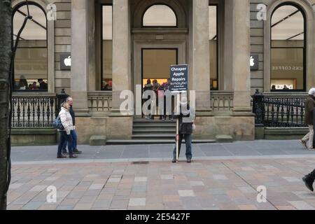 Buchanan Street, Glasgow, Schottland, Großbritannien. Ein Mann steht vor dem Apple-Einzelhandelsgeschäft mit einem Plakat, das billigere Apple-Reparaturen wirbt Stockfoto