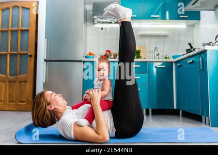 Eine Mutter mit ihrem Baby macht Fitness auf der Matte in der Küche und hebt ihre Beine. Seitenansicht. Das Konzept des Heimsporttrainings mit Kindern. Stockfoto
