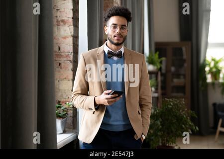 Porträt des jungen Geschäftsmannes in stilvoller Kleidung lächelnd in der Kamera Während der Verwendung seines Mobiltelefons im Büro Stockfoto