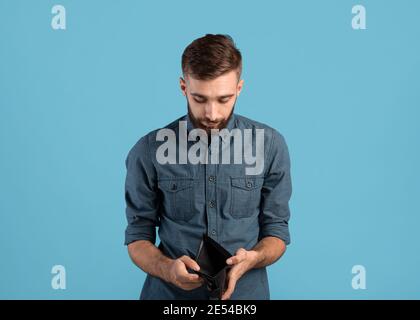 Armut und Arbeitslosigkeit. Verzweifelter junger Mann zeigt leere Brieftasche ohne Geld auf blauem Studiohintergrund Stockfoto