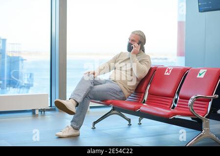 Reifer Mann in Maske sitzt auf dem Stuhl und spricht weiter Handy im Wartezimmer des Flughafens während einer Pandemie Stockfoto