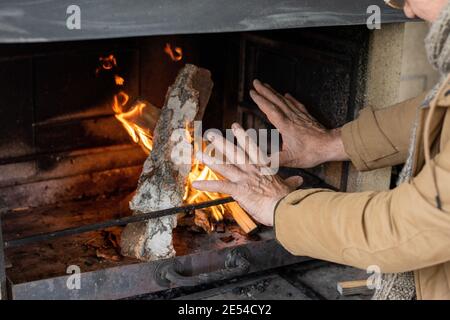 Hände eines älteren Mannes in brauner Jacke, der am Kamin steht Mit brennenden Feuerhölzern, während es an einem kühlen Herbsttag warm wird Im Landhaus Stockfoto