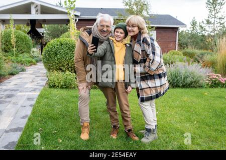 Glücklicher Youngster in Casualwear und seine liebevollen Großeltern suchen in Smartphone-Kamera mit Lächeln während der Herstellung Selfie Stockfoto