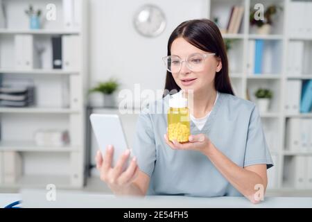 Junge lächelnde Ärztin in Uniform und Brille hält Smartphone Und zeigt dem Online-Patienten eine Flasche mit neuer Medizin Stockfoto