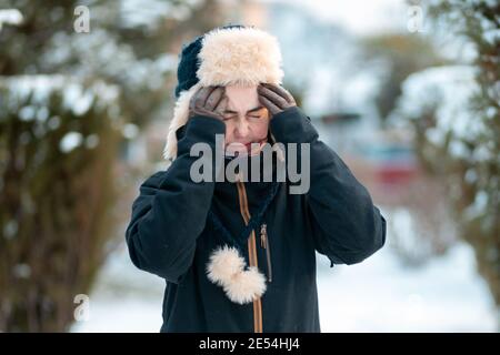 Porträt einer jungen, unzufriedenen kaukasischen Frau in einem Hut mit Ohrenklappen und einer Jacke, die ihre Stirn kratzt. Wintersaison, Schneefall. Stockfoto