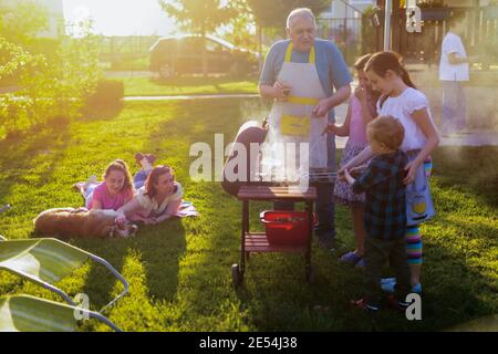 Stolzer, fröhlicher Großvater, der seinen Enkeln beibringt, wie man ein Barbecue macht. Spaß im Hinterhof am Sommertag. Stockfoto