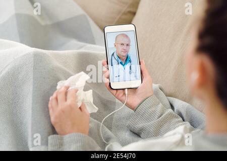 Junge kranke Frau mit Taschentuch und Smartphone mit reifen Mann Arzt auf dem Bildschirm sitzt auf der Couch und hört medizinische Beratung Stockfoto